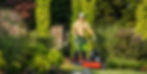 Man in cap and headphones mows a lush garden lawn on a red mower, surrounded by green shrubs and trees.