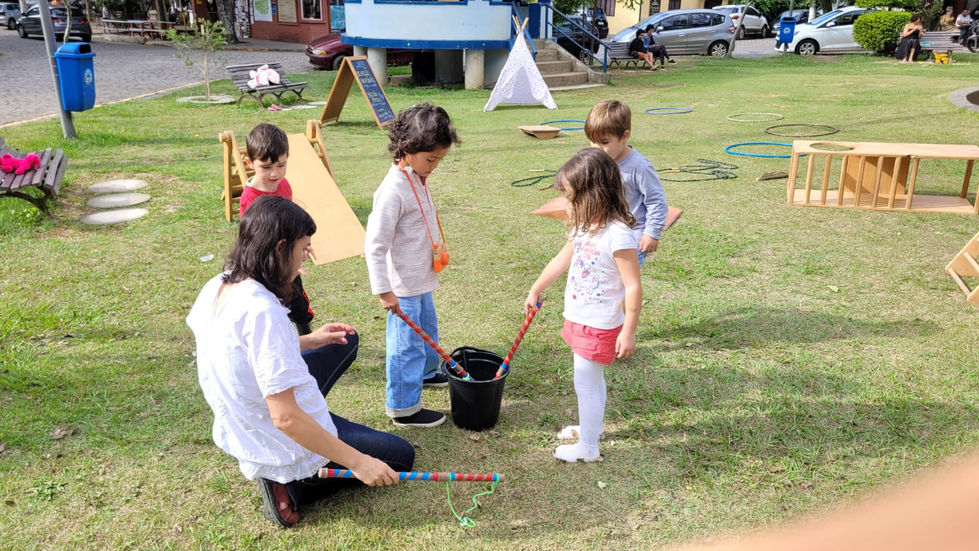 Crianças brincam ao ar livre com balde e bastões.