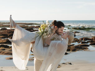 A bride and groom kiss on a beach in California, while the groom carries the bride in his arms and dips her down.