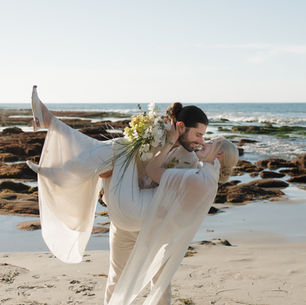 A bride and groom kiss on a beach in California, while the groom carries the bride in his arms and dips her down.