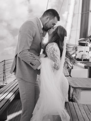 A bride and groom kiss in a black and white photo on a sailboat.