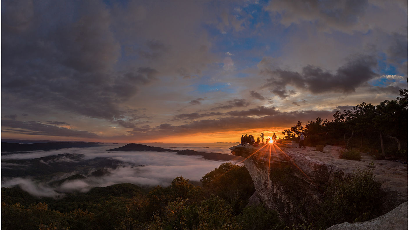 A group of people watch the sunrise from a dramatic rocky overlook above a valley surrounded by mountains.