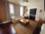 Open spacious living area with large windows, couch and armchair. there is a coffee table in the middle of the room and in the background the bedroom can be viewed through the doorway.