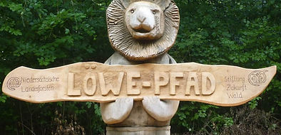 Hölzener Löwe mit einem Schild in den Pfoten auf dem "Löwepfad" steht.