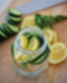 Jar of water with lemon, cucumber slices, and mint on a wooden surface. Knife and fresh slices in background. Refreshing mood.