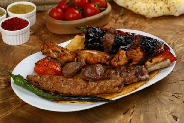 Bir tabakta karışık ızgara etler, közlenmiş biber ve domates. Arkada domatesler, baharat kapları ve lavaş ekmeği. Rustik ortam.