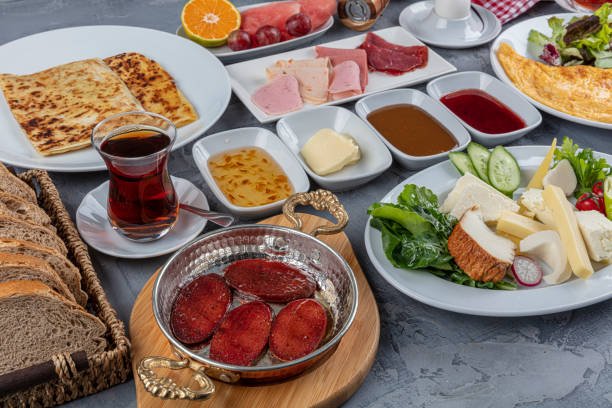 Türk kahvaltı sofrası: sucuklu tava, peynirler, zeytin, omlet, kanepede çay bardağı; fon gri, masa düzenli ve renkli.