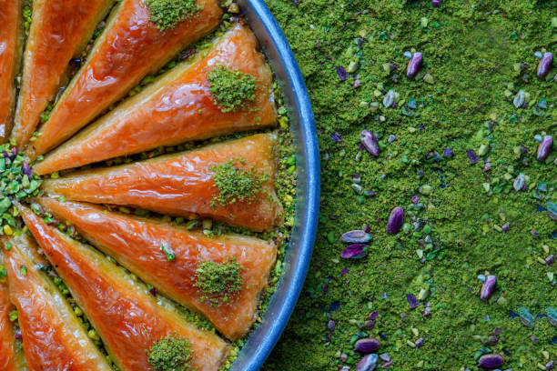 Tabağın içinde dilimlenmiş baklava yer alıyor. Üzeri fıstıkla kaplı. Arka plan yeşil fıstıklarla dolu. Canlı renkler dikkat çekiyor.