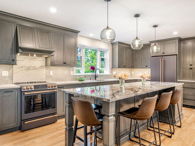 A large kitchen island surrounded by 4 brown leather high top chairs. The cabinetry in the background is medium grey and there is a window above the sink, showing greenery. This kitchen is in Littleton, Massachusetts and is newly remodeled.