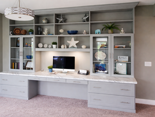 Custom built-in home office storage system with white cabinets, marble countertop desk, glass display shelves, and organized workspace design.