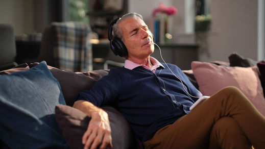 A man sits comfortably at home using the Lenire® tinnitus treatment device. He wears wireless Bluetooth headphones and has the Tonguetip® device placed in his mouth, connected to a small handheld controller. He appears relaxed and focused during the session.