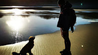 sagome di un cane e un donna sulla spiaggia di un lago con colline, il cielo e il sole sullo sfondo