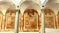Un porticato ad archi con antichi affreschi religiosi alle pareti e vasi di fiori sui muretti.