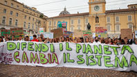 Manifestazione di Fridays for Future in una piazza italiana. I giovani sorreggono un grande striscione con la scritta verde e viola "DIFENDI IL PIANETA CAMBIA IL SISTEMA". Sullo sfondo un campanile.