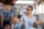 Two women in a discussion at an office, one gesturing with open hands. Background has large windows. One wears a striped shirt. Serious mood.