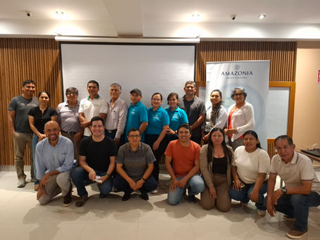 A group of seventeen participants pose for a photo in a conference room during an Amazonia Impact Ventures workshop, standing and kneeling in front of a projection screen and an AIV banner.