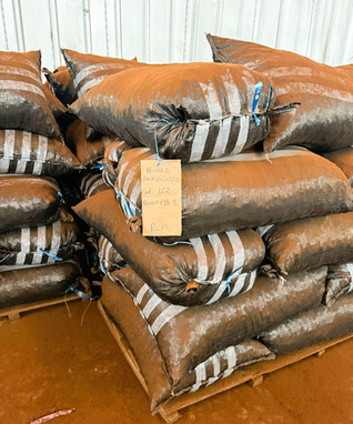Stacks of large brown sacks tied with blue string sit on pallets in a warehouse, with a handwritten label showing date, content and cooperative information.