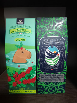 Two Sanchirio Palomar coffee packages placed side by side, one with a dark green design featuring a stylised bird and the other with a capybara illustration surrounded by coffee plants, highlighting Peruvian origin and varieties.