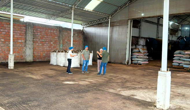 Four people wearing hairnets stand inside a warehouse discussing operations, surrounded by large bulk bags and stacks of smaller sacks in an agricultural processing environment.