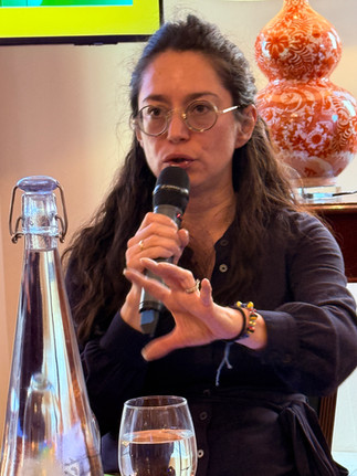 A woman speaking into a microphone during a discussion, with bottles of water and event materials on the table in front of her.