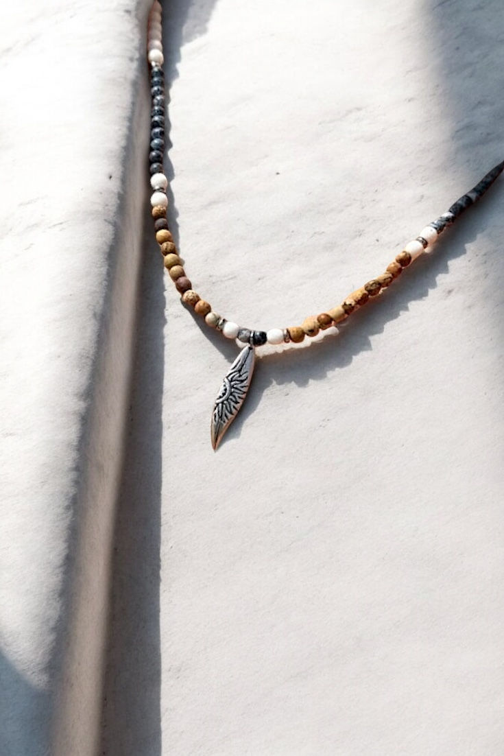 שרשרת חרוזים בגווני חום לבן וכחול כהה עם תליון בצורת גלשן מכסף שעליו יש חריטה של שמש מונחת על אבן בהירה 