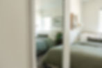 Bedroom with green bedding reflected in a decorative mirror. Art on walls, plant on side table. Soft, serene ambiance.