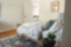 Cozy bedroom with a neatly made bed, grey pillows, and a white blanket. A potted plant on a side table, patterned rug, and a door ajar.