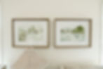 Two framed watercolor landscapes hang on a light wall. Below is a woven lamp shade. The setting is calm and minimalistic.