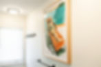 Framed artwork of a person lounging on an orange chair. Bright teal and orange colors. Minimal room with hooks, a window, and soft lighting.