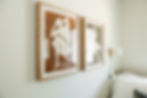 Two framed abstract art pieces of seated figures on a wall, with beige and brown tones. A bed and wall lamp are partially visible.