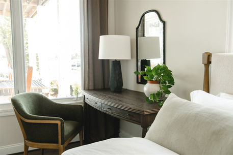 Cozy bedroom with a wooden desk, green chair, lamp, and potted plant. A mirror reflects light, and a neatly made bed is nearby. Calm ambiance.