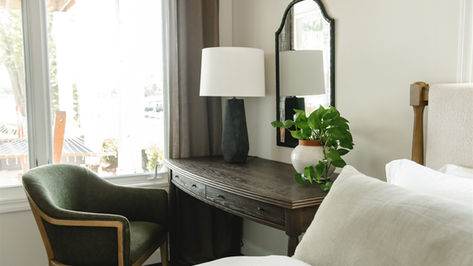 Cozy bedroom with a wooden desk, green chair, lamp, and potted plant. A mirror reflects light, and a neatly made bed is nearby. Calm ambiance.