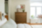 Wooden dresser with lamp and plant sits in a cozy bedroom with a large window and a white bed. Floral art hangs on the wall. Light and airy.