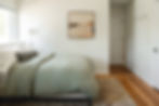 A cozy bedroom with a green bedspread, white wall, abstract painting, plaid pillow, and soft lighting. Wooden floor and hooks on the wall.