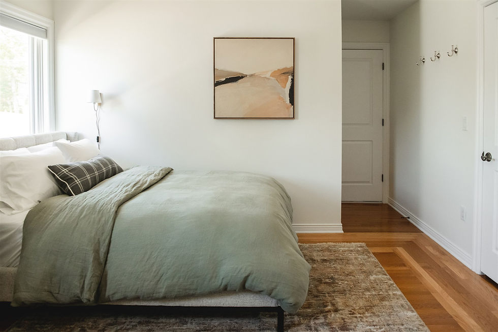A cozy bedroom with a green bedspread, white wall, abstract painting, plaid pillow, and soft lighting. Wooden floor and hooks on the wall.