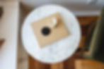 Marble table with a beige book, black-lidded white candle. Brown wooden floor, green chair nearby. Cozy, minimalist setting.
