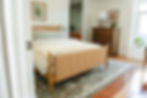Cozy bedroom with a wooden bed, beige blanket, large patterned rug, vibrant botanical art, wooden dresser, and a potted plant by the window.