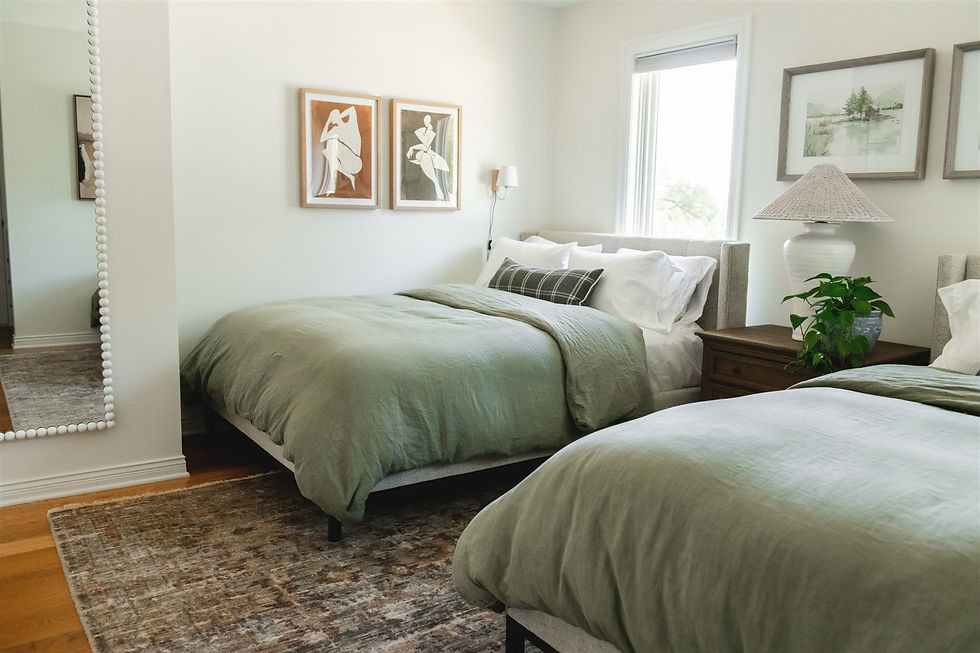 Two beds with green linens in a cozy bedroom, art on walls, a large mirror, wooden nightstand with plant, and window with natural light.