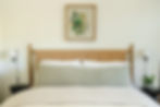 Beige bed with green cushion and white pillows, framed botanical art above. Two wall lamps and a small plant on dark side tables. Cozy bedroom.