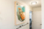 Framed painting of a person lounging in sunglasses on an orange chair. White walls, black towel rails, and a door in the background.