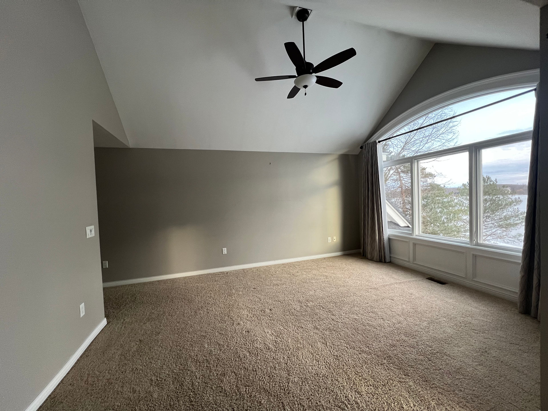 Spacious primary bedroom with beige carpet, large arched window, grey curtains, fireplace, ceiling fan, and glass doors showing a snowy landscape.