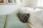 Bed with green duvet, white pillows, and a plaid black bolster pillow. Art on a white wall, soft light, and a cozy atmosphere.
