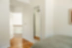 Hallway with a tall mirror on the right wall, wooden floor, open door leading to a carpeted area with railing. Bright, calm ambiance.