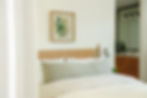 Cozy bedroom with a wooden headboard, green pillow, and white bedding. Botanical art on the wall, visible bathroom sink with wood cabinets.