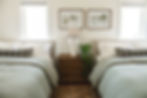 Two beds with gray plaid pillows and green duvets, flank a wooden nightstand with a white lamp and plant. Landscape art hangs above. Cozy mood.
