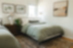 Cozy bedroom with a bed covered in green bedding, a wood nightstand with a potted plant, neutral art on walls, and a soft rug.