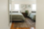 Bedroom with two beds, green comforters, checkered pillows, a lamp on a nightstand, plants, and framed art. Hardwood floor and a rug.