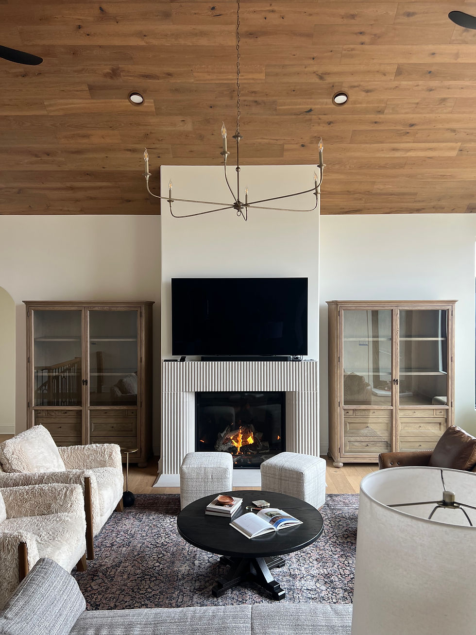 Modern living room designed by Lauren Figueroa with a wood ceiling, chandelier, and fireplace. Cream furniture and a round table holding books are on a patterned rug.