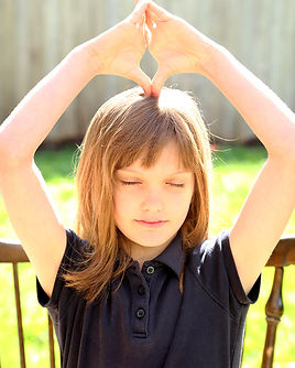 A young girl about 8 years old sits outside in a chair. Her hands are over her head and her fingertips are together and her two thumbs rest on top of her head. She is smiling slightly and her eyes are closed.