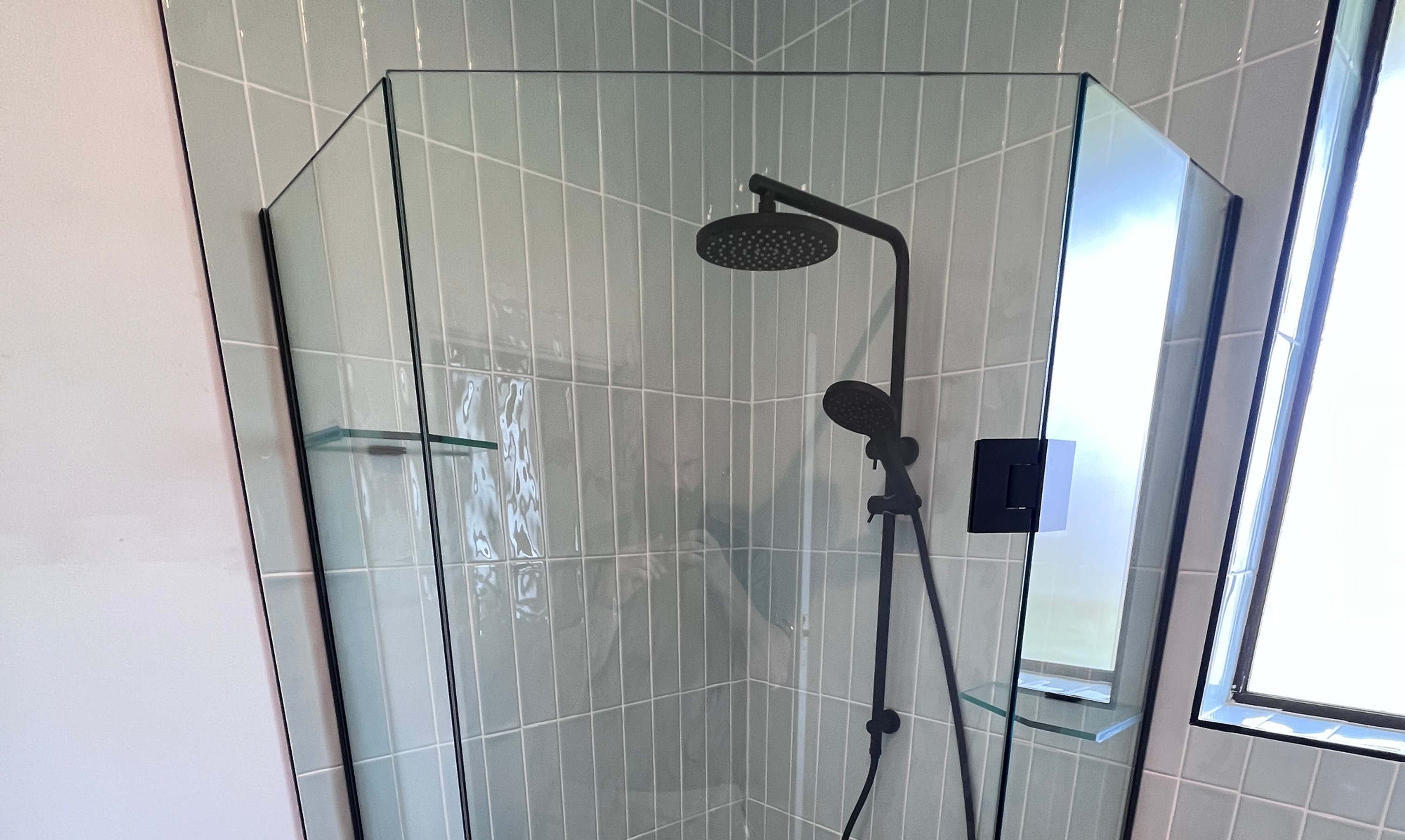 Modern bathroom shower with black fixtures, glass enclosure, pale green tile, wood vanity, and bright window light. Leaking shower Melbourne.